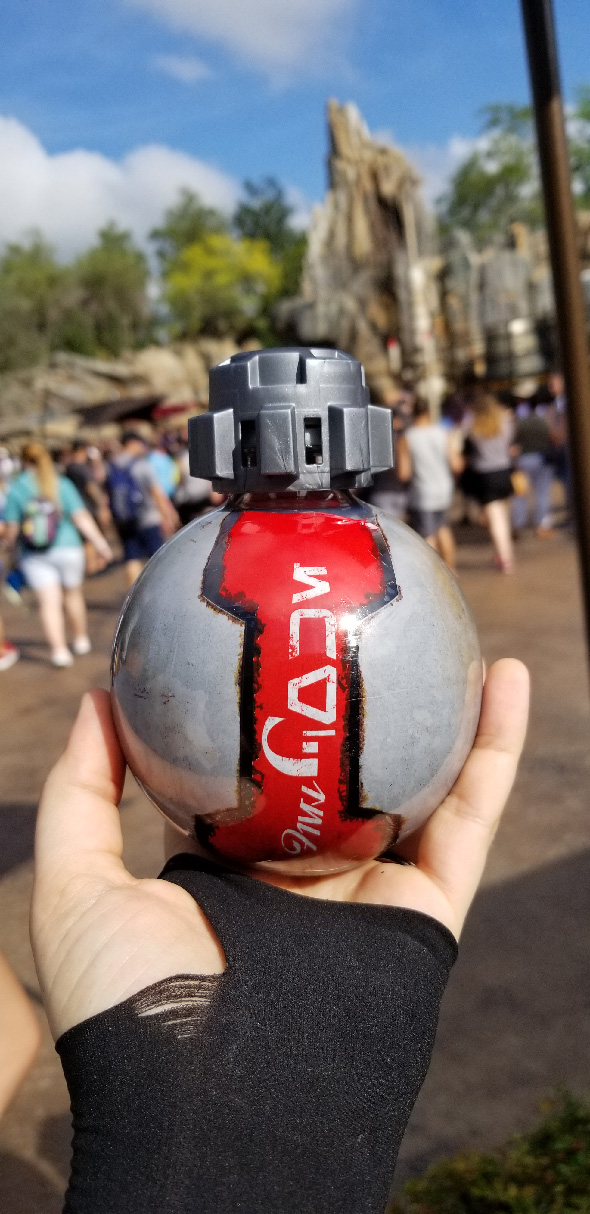 Gloved hand holding thermal detonator soda in front of spires and crowds